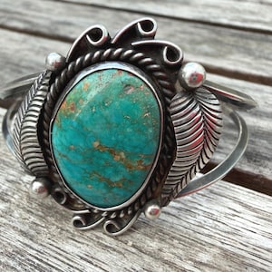 May include: A silver cuff bracelet featuring a large, oval-shaped turquoise stone with brown matrix veining. The centrepiece is framed by a twisted rope border, flanked by two detailed silver leaf motifs, and accented with small silver spheres and scrolls.