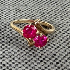May include: A gold-toned ring featuring two round, vibrant pinkish-red gemstones set in a bypass style. The band is smooth and metallic, with a visible hallmark stamped on the inner surface of the shank, showcasing a classic design with dual stones.