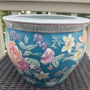 May include: A round ceramic planter with a vibrant teal background featuring a hand-painted floral pattern of pink peonies, yellow daffodils, and small purple blossoms. The rim is white with a thin black vine motif and a red accent line.