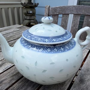 May include: A white ceramic teapot featuring a blue and white geometric patterned band around the lid and upper body. The teapot has a curved spout, a looped handle, and a lid with a small knob, decorated with subtle light blue leaf-shaped accents.