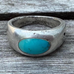 May include: A wide silver band ring featuring a smooth, polished finish and a single, oval-shaped turquoise gemstone set in the center. The metal surface displays a slightly weathered texture with visible scratches and natural signs of wear.