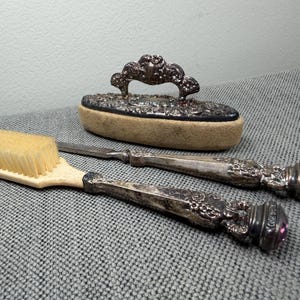 Könnte beinhalten: Eine ovale Kleiderbürste mit einem verzierten Silbergriff und einem beigen Borstenpolster, begleitet von einer langstieligen Bürste und einem Metallwerkzeug, beide mit komplexen silbernen Blumenmustern und kleinen violetten Steinen an den Griffenden.