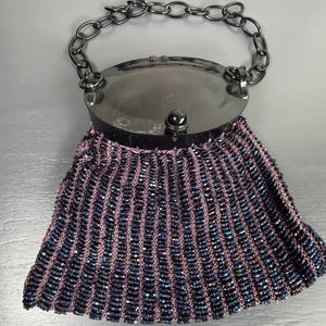 May include: A vintage beaded handbag with a silver-tone metal frame and chain handle. The bag features a dark purple and pink beaded design. The clasp is a small, round, black button. The bag is a small, elegant accessory.