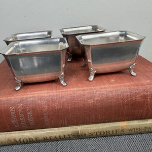 May include: Four small, silver-colored metal bowls with ornate feet are arranged on a vintage book with a red cover. The bowls have a square shape with slightly flared sides. The book's title is partially visible.