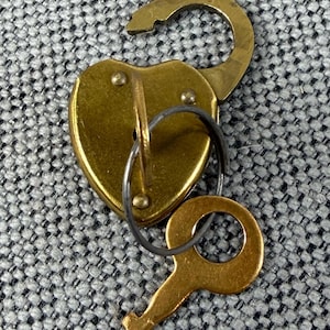 May include: A heart-shaped, open brass padlock with a matching key. The key is attached to the lock with a small metal ring. The lock is on a textured gray surface.