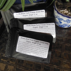 May include: Three black pouches with white labels that read "Shady Grove Farm Freeze-Dried Raw Organic Ginger 2 oz". The labels contain information about the product, including that it is a raw product made from USDA Organic Whole Ginger Root. The pouches are sitting on a brown woven surface.