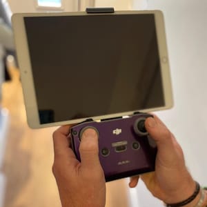May include: A purple drone controller with a tablet attached. The tablet is held in place by a black clip. The controller has two joysticks and several buttons. The tablet screen is black.