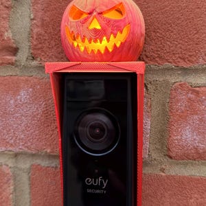 May include: A black Eufy Security video doorbell mounted with a red 3D-printed protective cover and a glowing orange jack-o'-lantern topper. The doorbell features a central camera lens, a blue circular light ring, and the Eufy Security logo.