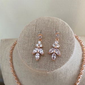 May include: A rose gold necklace and earrings set with clear cubic zirconia stones. The necklace is a delicate chain with small stones, and the earrings are drop earrings with a cluster of stones.