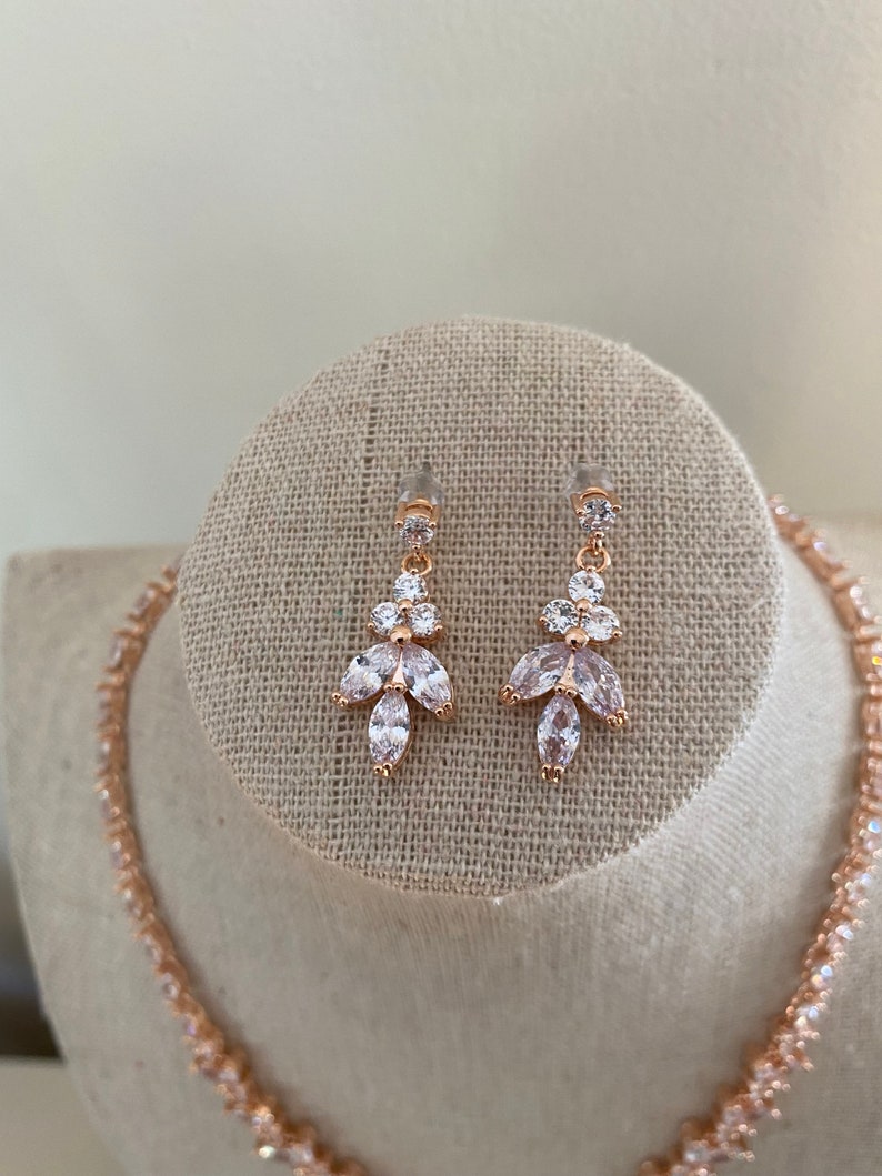 May include: A rose gold necklace and earrings set with clear cubic zirconia stones. The necklace is a delicate chain with small stones, and the earrings are drop earrings with a cluster of stones.