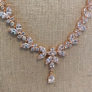 May include: A rose gold necklace with a teardrop pendant and a delicate floral design. The necklace is adorned with clear cubic zirconia stones.