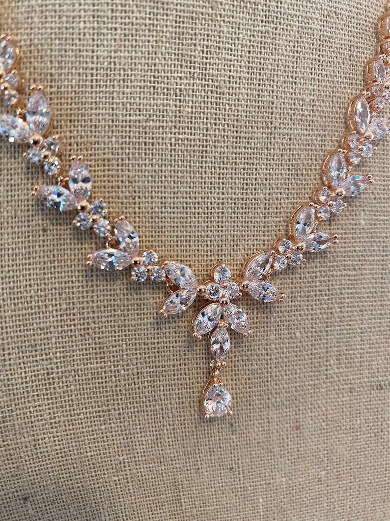 May include: A rose gold necklace with a teardrop pendant and a delicate floral design. The necklace is adorned with clear cubic zirconia stones.