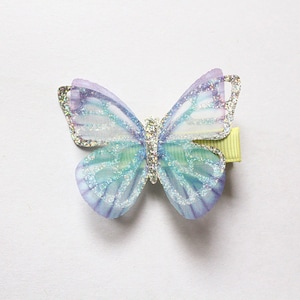 Pu&ograve; includere: Un fermaglio per capelli a forma di farfalla blu chiaro e viola con accenti di glitter. Il fermaglio &egrave; realizzato in tessuto trasparente e ha una clip a coccodrillo verde.