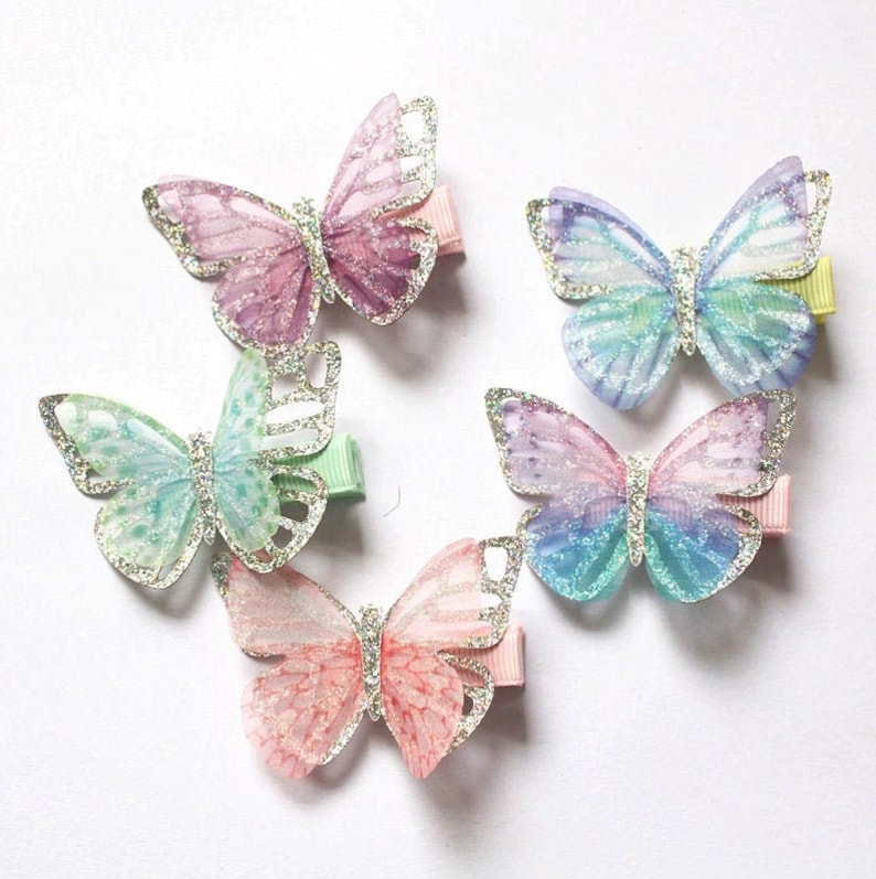 Pu&ograve; includere: Cinque fermagli per capelli a forma di farfalla in vari colori pastello con dettagli glitter. Le farfalle sono realizzate in un tessuto trasparente con un rivestimento di glitter argentato.