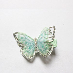 Pu&ograve; includere: Un fermaglio per capelli a forma di farfalla verde con glitter e contorno argentato.