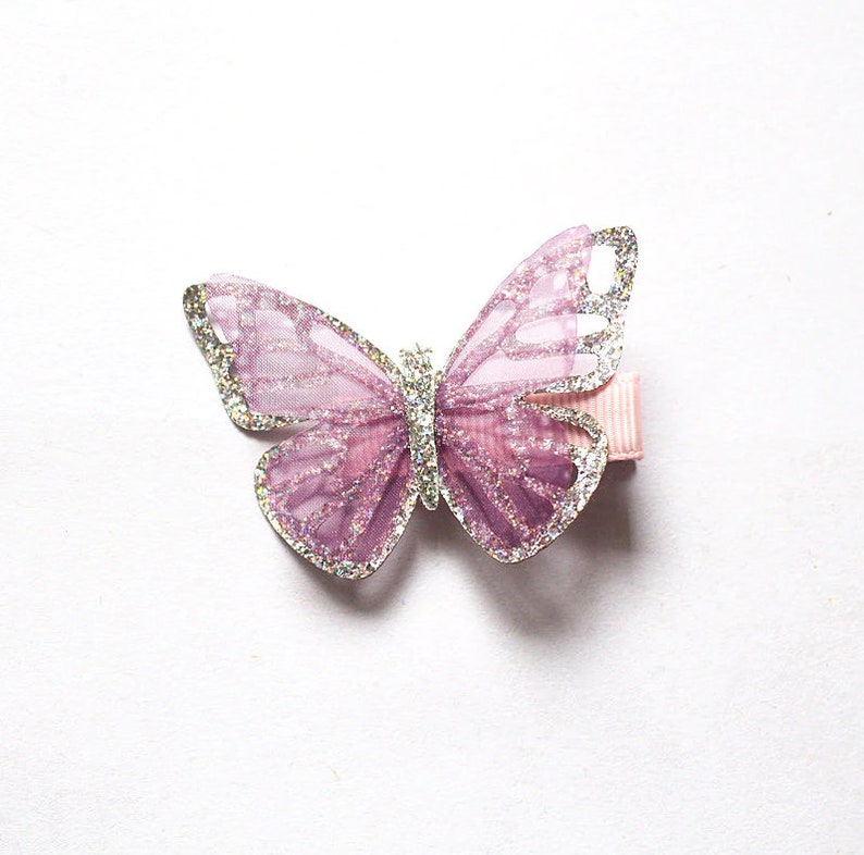 Pu&ograve; includere: Un fermaglio per capelli a forma di farfalla viola con accenti di glitter argentati. Il fermaglio &egrave; realizzato in un tessuto morbido e trasparente e ha un nastro rosa.