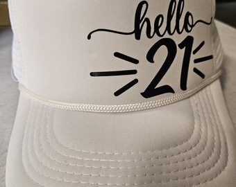 Puede incluir: Gorra de camionero blanca con visera curva y parte trasera de malla. La gorra presenta el texto "hello 21" en escritura negra y letras de bloque, con líneas estilizadas. Un detalle de cuerda blanca acentúa la visera.