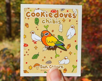 May include: A yellow and orange Sun Conure bird with a white background. The background has a pattern of leaves, mushrooms, and acorns. The text "Cookie'doves chibis" is at the top of the card. The text "Sun Conure Aratinga solstitialis" is at the bottom of the card.