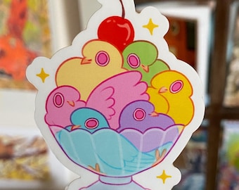 Puede incluir: Una pegatina colorida de un plato de postre de vidrio lleno de pájaros de colores pastel y una cereza roja en la parte superior. Los pájaros están dispuestos en un patrón circular y tienen ojos pequeños y redondos. La pegatina tiene un efecto brillante y brillante.