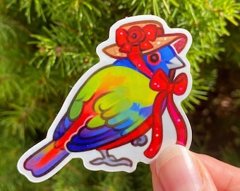 Puede incluir: Una pegatina colorida de un pájaro que lleva un sombrero de paja con un lazo rojo. El pájaro tiene la cabeza azul, el cuerpo verde, las alas amarillas y las plumas de la cola rojas.