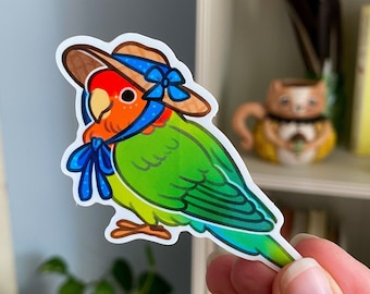 Puede incluir: Un agapornis verde y naranja con un sombrero de paja y una cinta azul. El pájaro está mirando hacia la izquierda y tiene la cabeza inclinada hacia abajo.