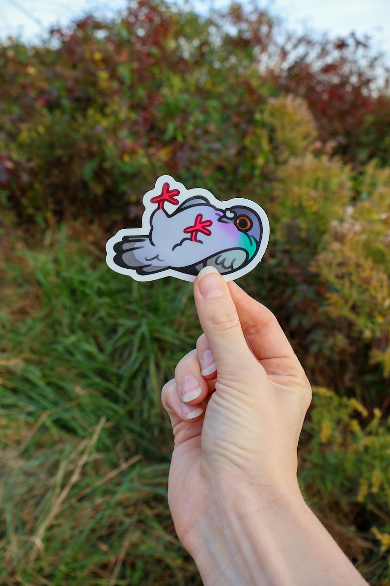 May include: A sticker of a cartoon pigeon with red feet, a gray body, and a purple and green head. The sticker is being held in a hand against a blurred background of green and brown foliage.