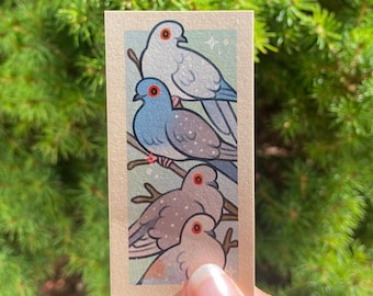 Puede incluir: Un primer plano de una pequeña pintura que representa cuatro palomas posadas en una rama. Las palomas están pintadas en tonos de azul, gris y blanco con ojos rojos. El fondo es de color azul claro con motas blancas.