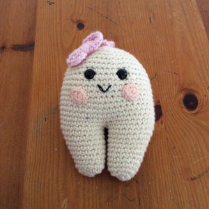 May include: A crocheted plush toy shaped like a tooth with a pink bow on top. The tooth has a smiling face with black eyes and pink cheeks.