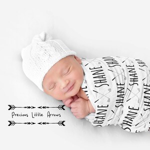 May include: A white swaddle blanket with black text that says "SHANE" repeated multiple times. The blanket is wrapped around a sleeping baby wearing a white knitted hat. The text "Precious Little Arrows" is printed below the baby in black.