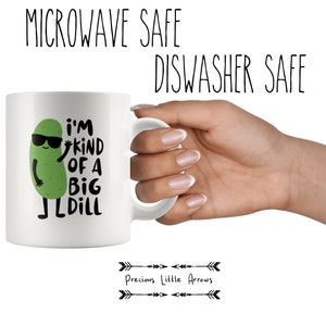 May include: White ceramic mug with a green cartoon pickle wearing sunglasses and saying "I'm kind of a big dill". The mug is microwave and dishwasher safe.