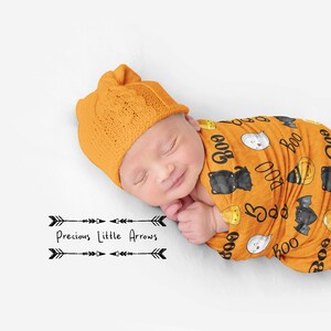 May include: A newborn baby swaddled in an orange and black Halloween-themed swaddle with a pattern of bats, ghosts, candy corn, and the word "Boo". The baby is wearing a matching orange knitted hat. The text "Precious Little Arrows" is printed below the baby.