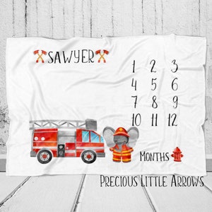 May include: A white baby milestone blanket with a red fire truck, a grey elephant dressed as a firefighter, and the text "SAWYER" and "PRECIOUS LITTLE ARROWS". The blanket has numbers 1-12 for each month of the year.
