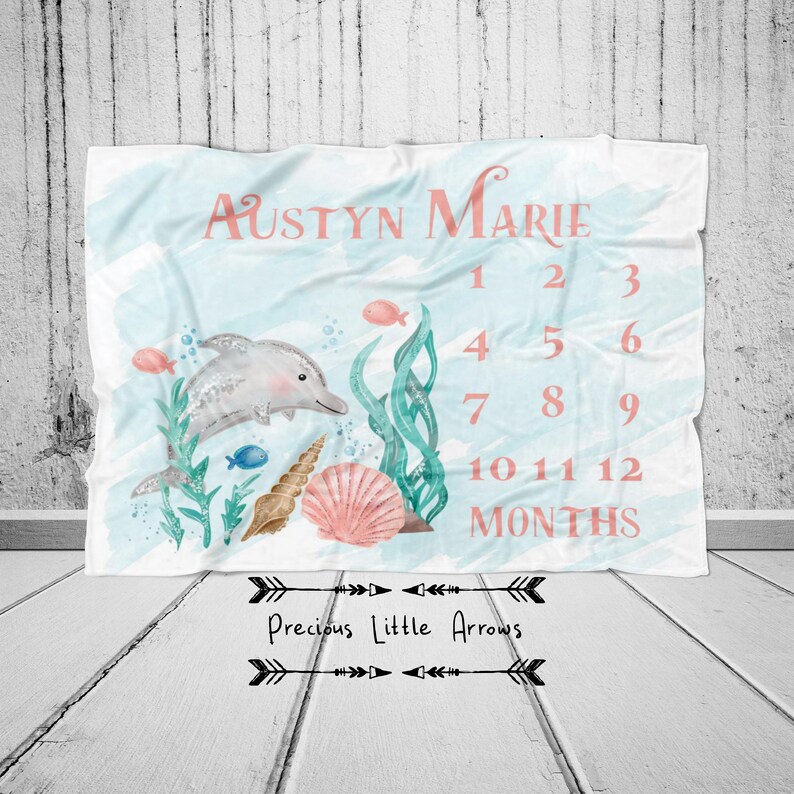 May include: A baby milestone blanket with a watercolor design featuring a dolphin, seashells, and aquatic plants. The blanket is light blue with the name "Austyn Marie" and numbers 1-12 months in coral.