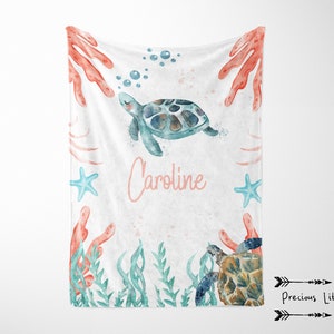 May include: A white baby blanket with a watercolor design featuring a sea turtle, coral, starfish, and aquatic plants. The name "Caroline" is written in pink script. The blanket has a soft, inviting appearance.
