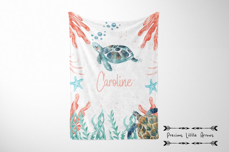 May include: A white baby blanket with a watercolor design featuring a sea turtle, coral, starfish, and aquatic plants. The name "Caroline" is written in pink script. The blanket has a soft, inviting appearance.