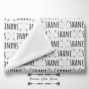 May include: White baby blanket with a repeating pattern of the name "Shane" and crossed golf clubs in black and gray.