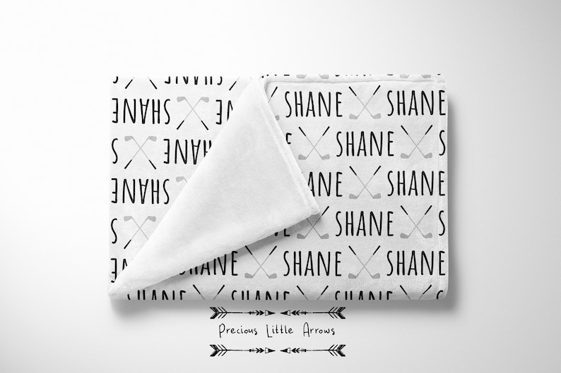 May include: White baby blanket with a repeating pattern of the name "Shane" and crossed golf clubs in black and gray.