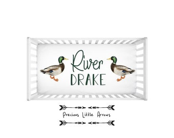 May include: White crib sheet with a green and brown duck illustration on each side and the text "River Drake" in green. The bottom of the sheet has a black arrow design with the text "Precious Little Arrows".