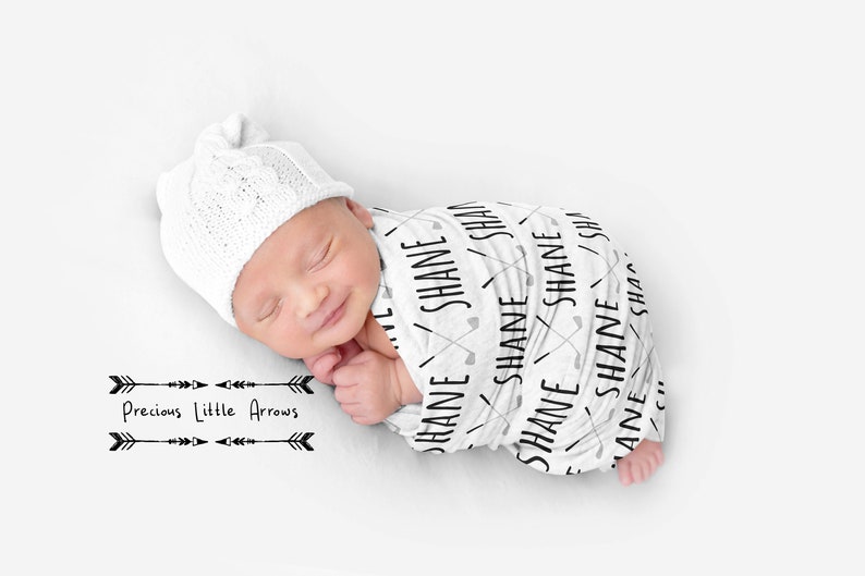 May include: A white swaddle blanket with black text that says "SHANE" repeated multiple times. The blanket is wrapped around a sleeping baby wearing a white knitted hat. The text "Precious Little Arrows" is printed below the baby in black.