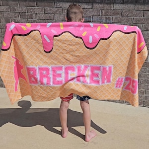 May include: A large beach towel with a pink ice cream cone design. The top of the towel has pink frosting with sprinkles. The towel has the name "BRECKEN" in pink block letters and the number "29". A pink silhouette of a baseball player is on the left side.
