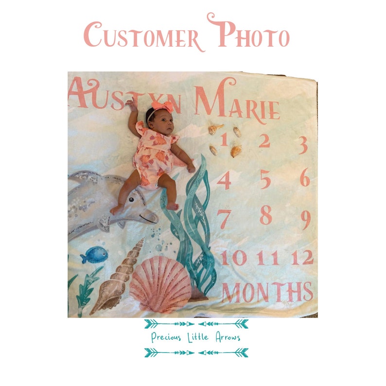 May include: A baby lies on a milestone blanket with an ocean theme. The blanket features a dolphin, seashells, and seaweed illustrations. The text "AUSTYN MARIE" and numbers 1-12 are printed on the blanket, along with the words "MONTHS" and "CUSTOMER PHOTO".
