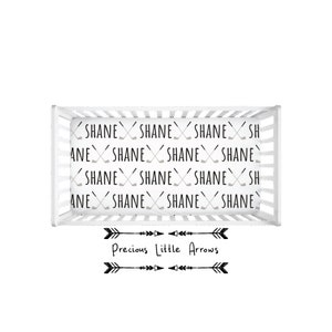 May include: A white crib with a fitted sheet featuring a repeating pattern of the name "Shane" and crossed hockey sticks. The crib is decorated with a black and white arrow design and the text "Precious Little Arrows".