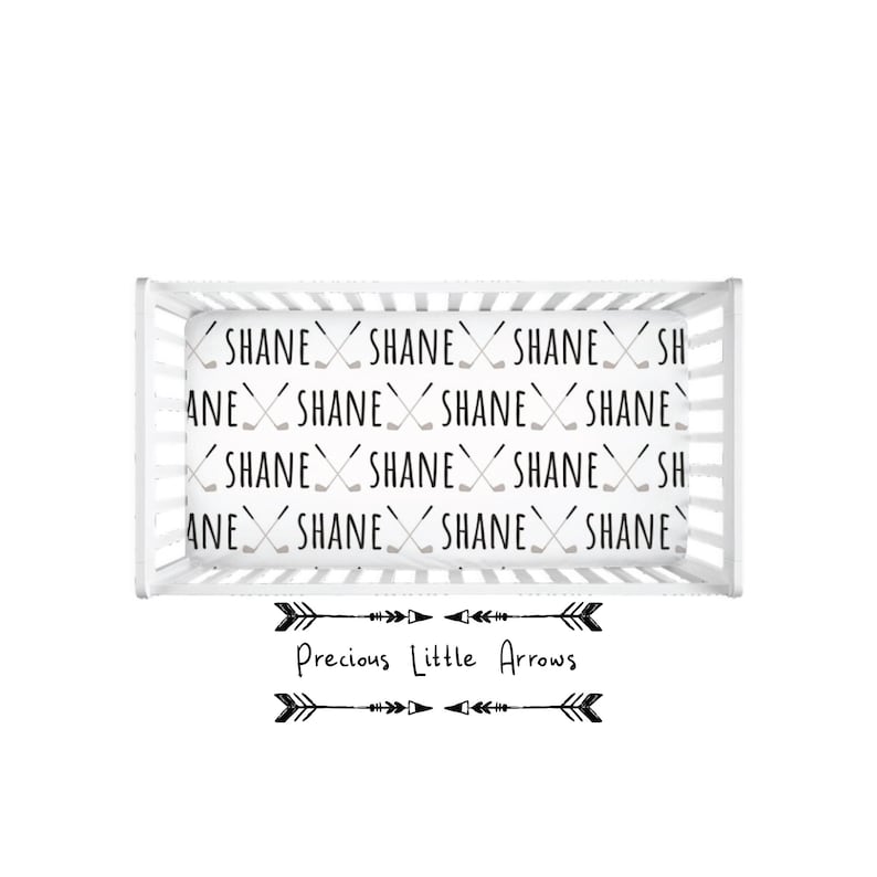 May include: A white crib with a fitted sheet featuring a repeating pattern of the name "Shane" and crossed hockey sticks. The crib is decorated with a black and white arrow design and the text "Precious Little Arrows".