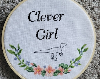 May include: White embroidery hoop with a floral border and the text "Clever Girl" in black lettering. A black silhouette of a velociraptor is in the center of the hoop.