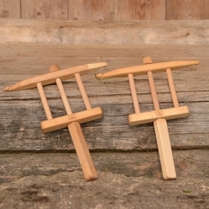 May include: Two wooden yarn swift tools, each with a T-shaped handle and a rectangular frame with vertical bars. The tools are light brown and rest on a weathered wooden surface, suggesting a rustic or handmade craft theme.