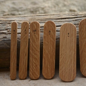 Può includere: Un set di sette strumenti in legno con la parte superiore arrotondata, ciascuno contrassegnato con un numero e una piccola freccia. Gli strumenti sono di varie dimensioni, da stretti a larghi, e sono realizzati in legno marrone chiaro. Gli strumenti sono disposti in fila.