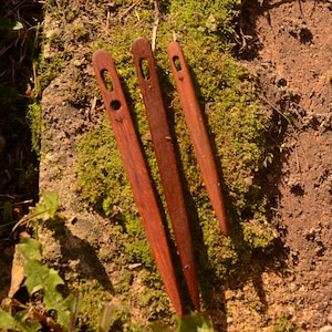 Op de afbeelding: Drie donkerbruine houten naalden met langwerpige ogen, naast elkaar geplaatst. Elke naald heeft een taps toelopend punt aan één uiteinde en een gladde, gepolijste afwerking, die de natuurlijke nerf en de diepe roodbruine kleur van het hout toont.