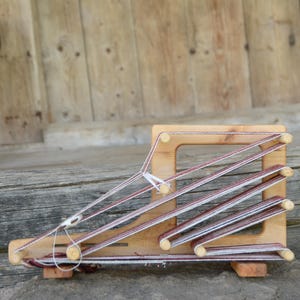 以下が含まれることがあります： 白い糸とバーガンディ色の糸がフレームに張られた、小さな木製の織機。織機は長方形で、中央に正方形の開口部があり、風化した木製の背景に配置されています。