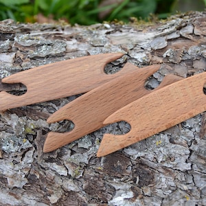 Puede incluir: Tres herramientas de tejido de madera, también conocidas como 'lanzaderas de tejido', están dispuestas sobre un trozo de corteza de árbol. Las herramientas están hechas de madera marrón claro y tienen un acabado liso y pulido.