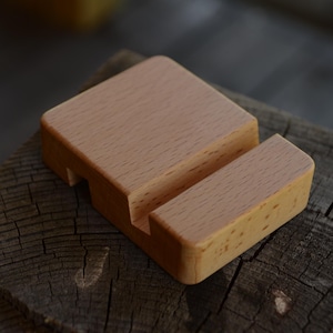 May include: A light-colored wooden phone stand. The stand is rectangular with a slot to hold a phone and a base. The wood grain is visible, and the stand is sitting on a weathered wooden surface. A simple, functional design.